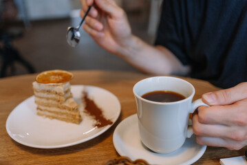 Close-up of man drinking coffee with slice of layer cake on wooden table. Delicious sweet dessert with dried orange slice and an invigorating americano drink. Snacking on fast carbohydrates.