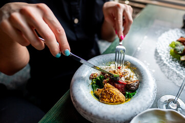 Close-up of a person dining at an elegant restaurant, focusing on their hands cutting a gourmet dish with a fork and knife. The table is set with fine cutlery and a glass of wine