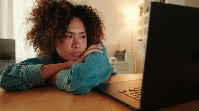 Bored young woman sitting at table in living room looking at laptop