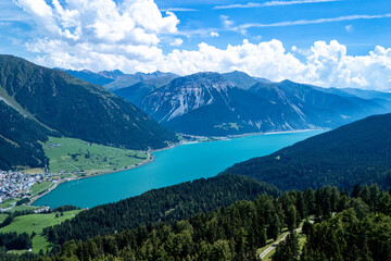 A panoramic view of turquoise lake surrounded by mountains and lush greenery on a sunny day in the Alps