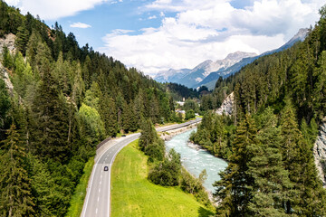 A scenic view of a winding road alongside a river surrounded by lush greenery and mountains on a sunny day in the Alps