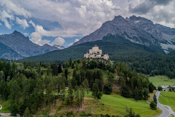 Aerial view of a castle perched on a hill surrounded by lush forests and towering mountains during a cloudy day