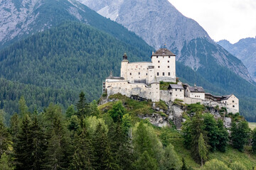 A majestic castle perched on a rocky hill surrounded by lush greenery and mountains in the background during a cloudy day