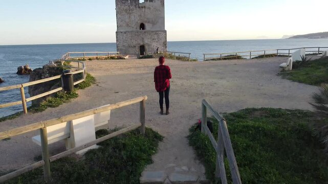 Aerial view of a middle-aged woman dressed in red walking towards a watchtower situated on a cliff on the coast of Casares in Malaga, Andalusian