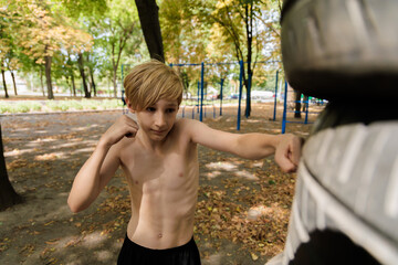 An athletically built teenager strikes with his hands on athletic equipment. Athletic teenage boy in the park outdoor workout session