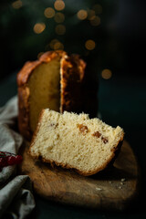 Sliced homemade Christmas panettone, made as holiday present with pine and red berries decorations on wooden table on rustic kitchen with Christmas lights on background.