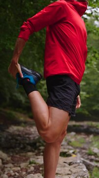 Unrecognisable young man stretching quadricep muscle in the forest. Athlete warming up in nature.