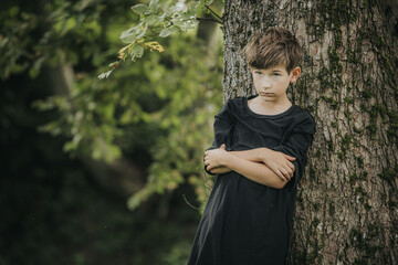 A young boy in a black shirt and khaki pants leans against a large tree, arms crossed, and gazes...