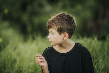 A young boy in a black shirt looks thoughtfully to the side while gently holding a blade of grass....