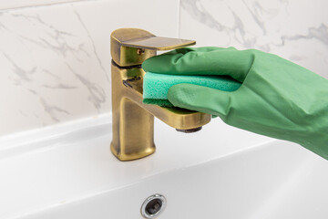 Man or woman in green rubber gloves cleans the gold faucet in bathroom with sponge