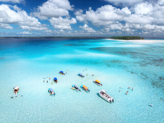 Aerial view of colorful boats in clear azure water in summer. Mnemba island, Zanzibar. Top view of sandbank in low tide, blue sea, white sand, swimming people, yachts, dramatic sky with clouds. Ocean