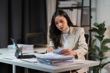 Late Night Data Dive: Focused Asian businesswoman analyzing financial reports at her desk, illuminated by the warm glow of her laptop in a contemporary office setting. 