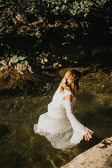 A woman in a white dress wades through a shallow, clear river surrounded by greenery and sunlight.