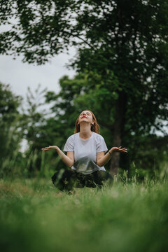 A young woman engages in mindfulness meditation outdoors, seated on grass in a tranquil park environment, surrounded by lush greenery.
