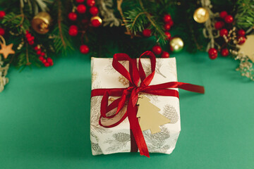 Merry Christmas! Stylish Christmas gift with red ribbon and green fir branches, red berries and gold ornaments on green background. Xmas background. Season's greetings