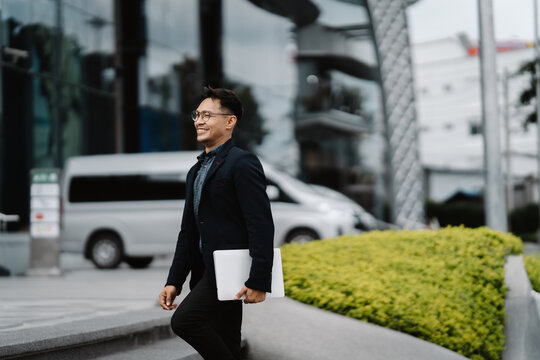Confident Stride: A young businessman exudes confidence and ambition as he confidently walks towards his future, laptop in hand. His stylish attire and bright smile suggest an optimistic and successfu