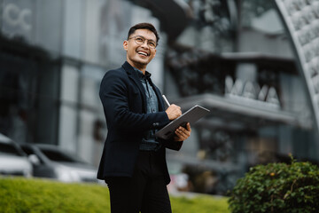Confident Businessman:  A young, successful Asian businessman in a suit and glasses confidently reviews a document on his tablet,  smiling with a forward-thinking attitude. He stands in an urban setti