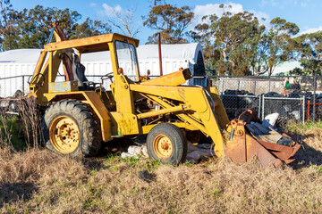 Photograph of an old and rusty industrial front end loader with a bucket full of rubbish that has been abandoned amongst other trash in a grassy field.