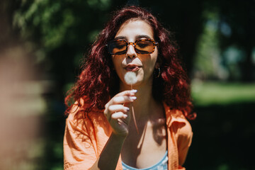 Young woman with red hair and sunglasses blowing dandelion seeds on a sunny day in the park. Joy...