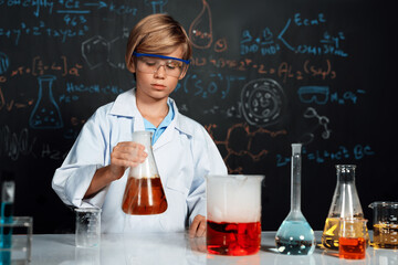 Blonde hair schoolboy in laboratory wear lab coat stand and learn science of chemistry technology in STEM class. The student hold beaker with orange liquid. On the table put many flask. Erudition.