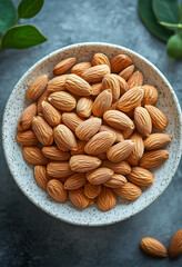 A bowl of fresh almonds on a textured surface with subtle greenery. A bowl filled with raw almonds rests atop a textured surface, surrounded by delicate greenery