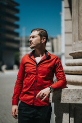 Confident young businessman standing outdoors in an urban setting. He is wearing a red shirt and looking contemplative, conveying professionalism and ambition.