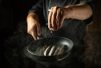 Adding salt to fish in a frying pan. Chef cooking capelin in the hotel kitchen. Concept on a dark background of preparing fish menu