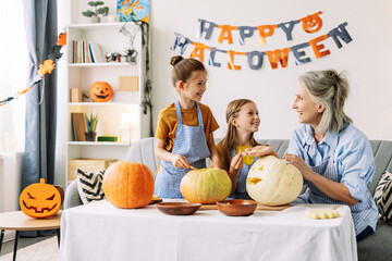 Smiling, happy grandmother and granddaughters carving pumpkins for halloween celebration at home