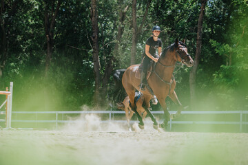 A young woman enjoying horseback riding in a sunny outdoor arena with lush green trees in the...