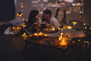 Romantic surprise for girlfriend or boyfriend. Bedroom prepared for watching old movies with popcorn, decorated with lights and candles. Cozy home Christmas atmosphere, hot chocolate with marshmallow