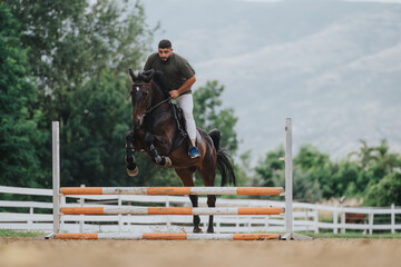 A man is riding a horse and skillfully jumping over an obstacle in an outdoor equestrian event, surrounded by nature.