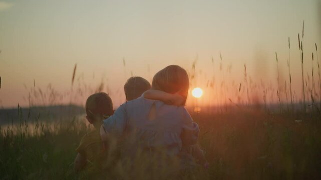 A back shot of a mother in a blue gown sitting in a field during sunset, holding her younger child on her lap while the older child sits beside her. They all gaze out at the serene lake