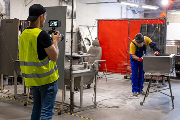 man in a yellow vest films a worker handling metal equipment inside an industrial workshop,...