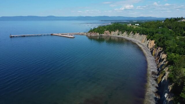 Vol de drone au-dessus d'une falaise rocheuse verdoyante formant le littoral d'une baie montagneuse accompagn&eacute; d'un vieux quai et d'une marina baignant sur l'eau calme. ( Baie-des-Chaleurs, Qu&eacute;bec )
