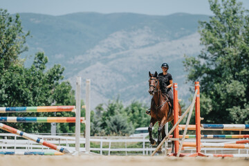 Horse rider jumping over colorful barriers during training in an outdoor arena with mountains in...