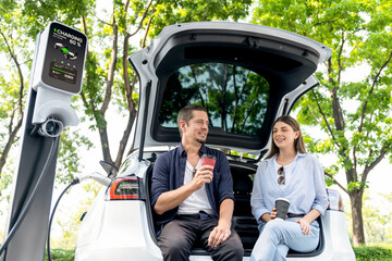 Obraz premium Lovely young couple drinking coffee while recharging battery for electric car during road trip travel EV car in natural forest or national park. Eco friendly travel during vacation and holiday. Exalt