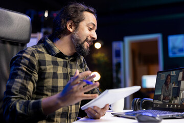 Closeup of young male university student communicating with his classmates on video call, comparing notes through laptop. Smiling man grasping notebook while having a virtual meeting.