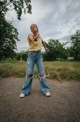 A young woman dressed in ripped jeans and a yellow shirt spends time outdoors, appreciating the serene natural environment under a cloudy sky.