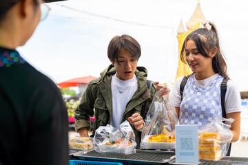 Young Asian man and woman buying and eating street food at railway station with using mobile app on smartphone scan QR code making online payment during travel local town on summer holiday vacation