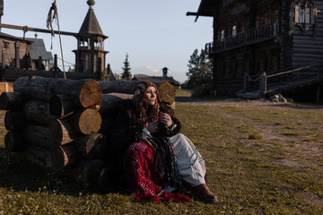 A shaman woman in a folk costume sits by a well, in a village among wooden architecture, Slavic or Nordic style