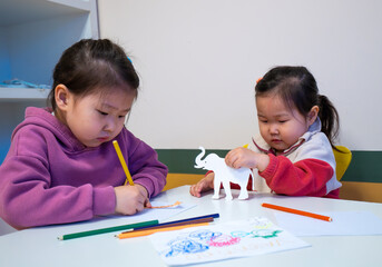 Two young girls are sitting at a table, drawing and coloring