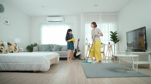 Caucasian mom and asian child doing housework together and dancing to music. Attractive mother and daughter cleaning modern bedroom while sing along and jump to relaxing music. Lifestyle. Pedagogy.