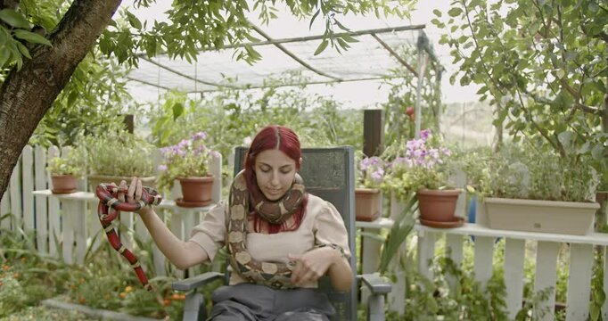 A woman sits outside in her garden and plays with her pet snakes, a red milk snake and a beige python.