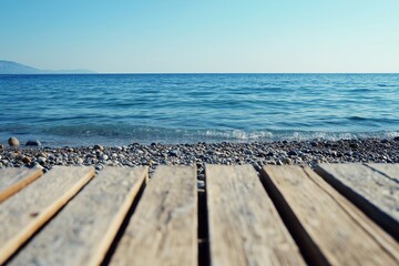 Fototapeta premium A tranquil seascape featuring a calm blue ocean meeting a pebbled shore, viewed from a wooden pier under a clear sky.