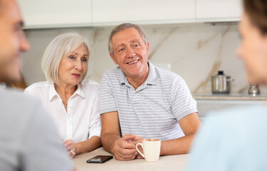 Obraz premium Smiling elderly parents and adult children sit at the table with tea and talk about home life