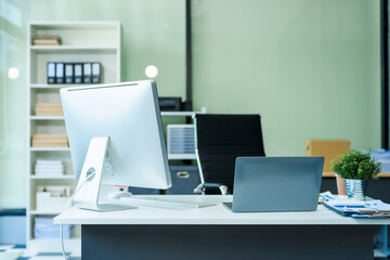 A sleek modern desk with a computer and laptop sits empty in a glass office, reflects a contemporary style with no people present, emphasizing a clean, professional environment.