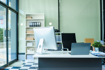 A sleek modern desk with a computer and laptop sits empty in a glass office, reflects a contemporary style with no people present, emphasizing a clean, professional environment.