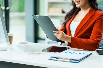 young Asian businesswoman in a formal suit works at her desk in a modern glass office, consulting on LGBTQ-inclusive strategies across various business domains, including HR, IT, and marketing.