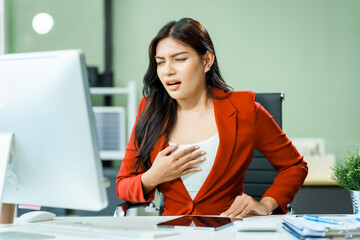 Chest pain of young Asian businesswoman in a formal suit experiences office syndrome and pain while working at desk in a modern glass office, consulting on diverse business topics