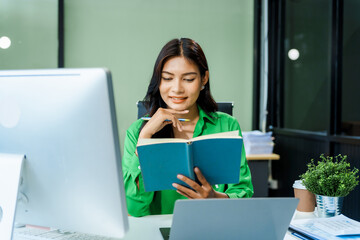 successful and happy young Asian businesswoman in a formal suit works at her desk in a modern glass...